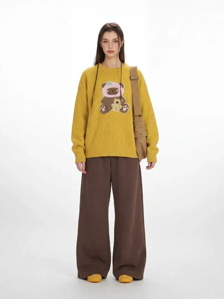 Woman wearing a yellow long-sleeved crew neck sweater with a teddy bear appliqué, paired with brown wide-leg pants and a tan crossbody bag.