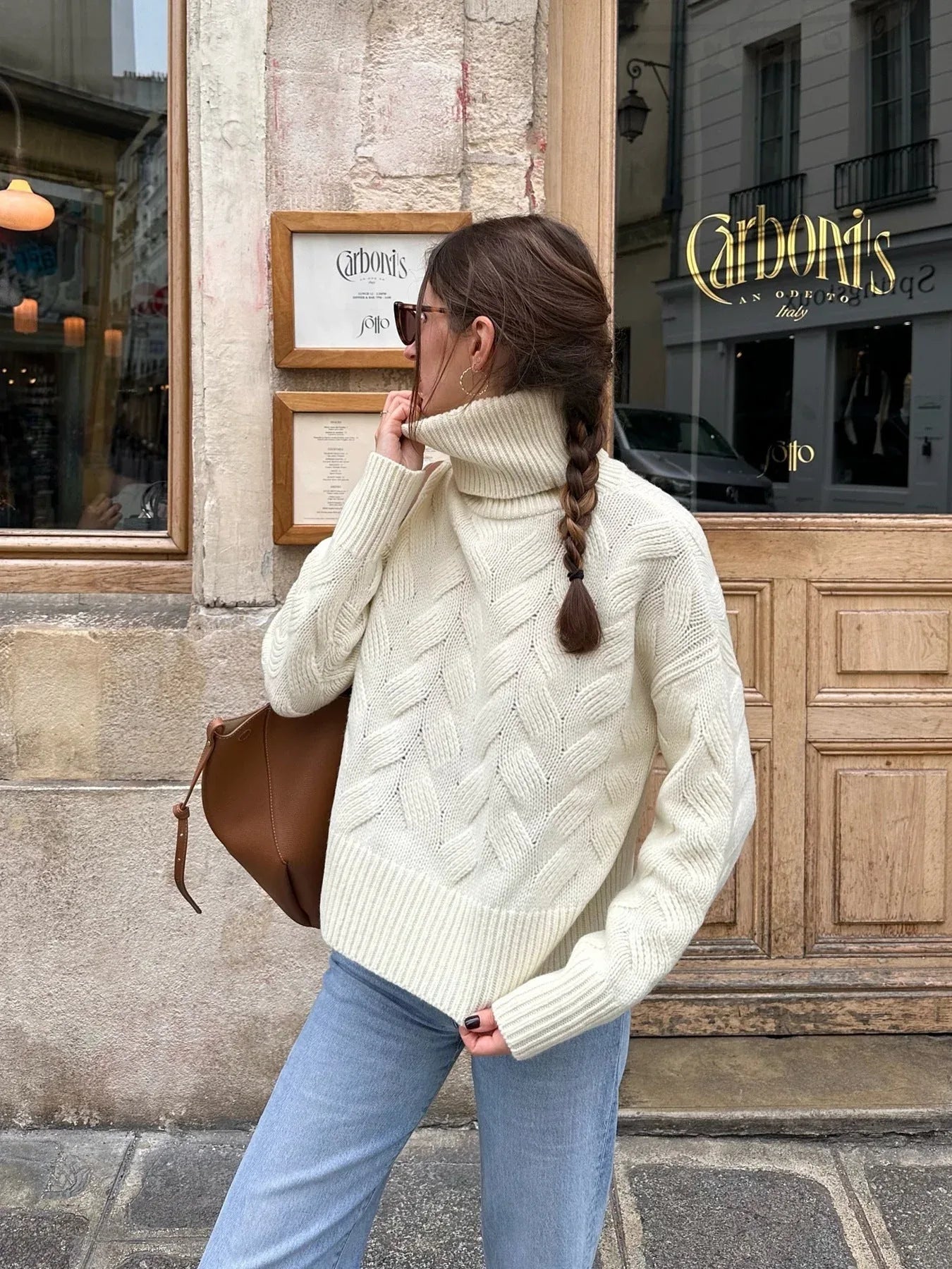 Woman with a braid wearing a cream cable knit turtleneck sweater and light wash jeans, posing outdoors.