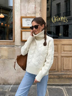 Close-up of a woman wearing a cream cable knit turtleneck sweater and light wash jeans, outdoors.