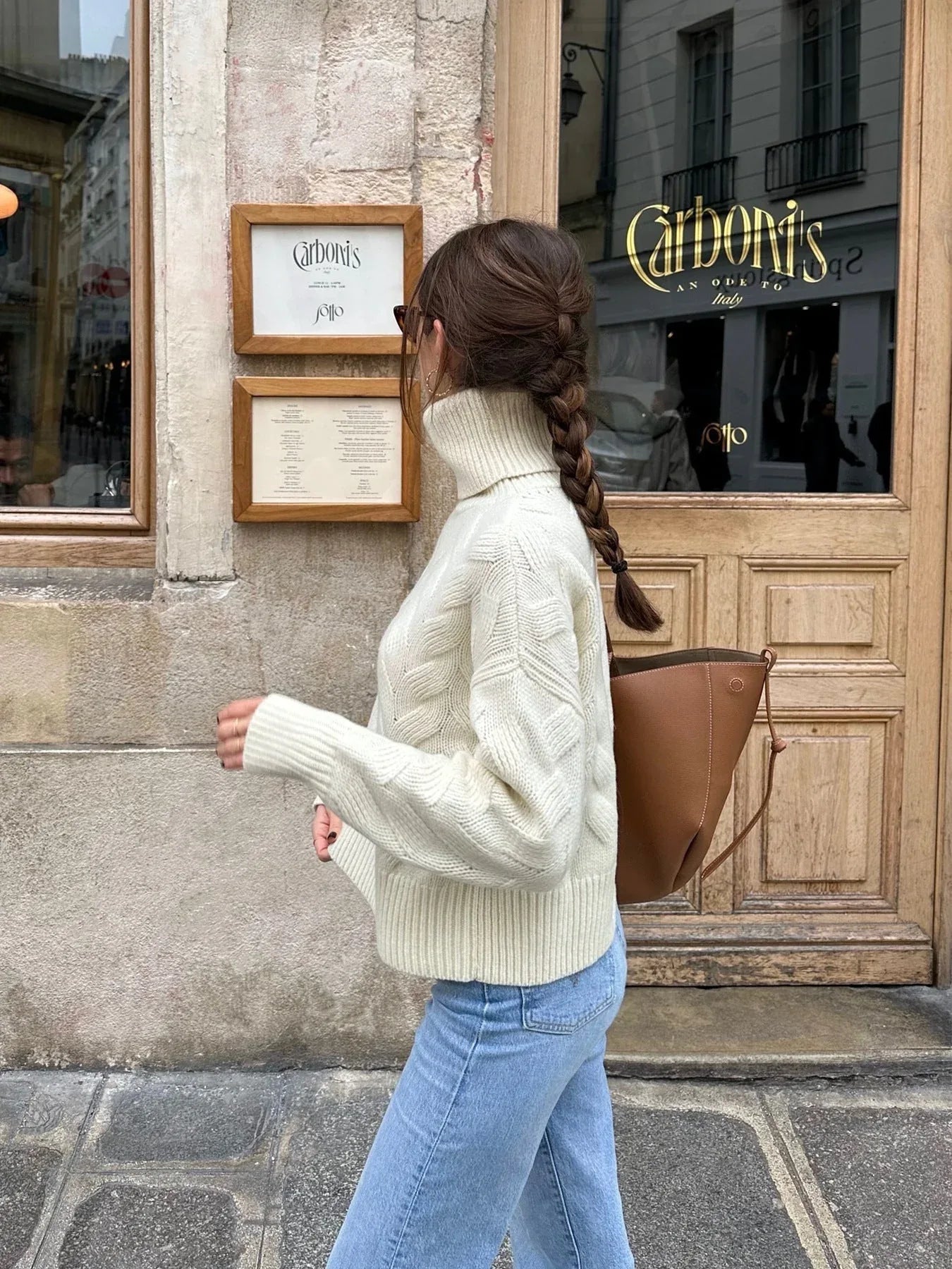 Side view of a woman wearing a cream cable knit turtleneck sweater and light wash jeans, walking outdoors.