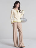 Full body shot of a woman in a cream long-sleeve henley shirt with lime green trim, beige trousers, and a silver handbag.