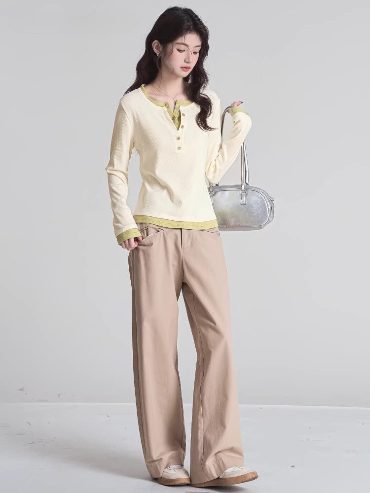 Full body shot of a woman in a cream long-sleeve henley shirt with lime green trim, beige trousers, and a silver handbag.