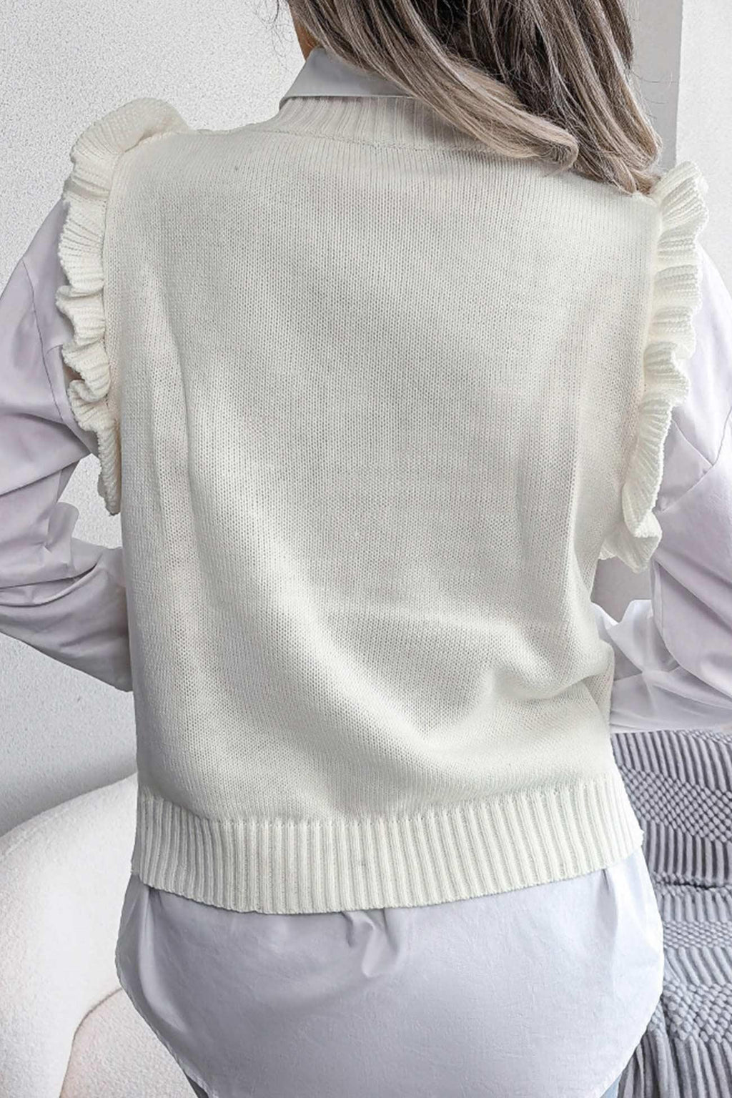 Back view of a woman wearing a white sleeveless knit vest with a diamond pattern and ruffle trim over a white button-down shirt.
