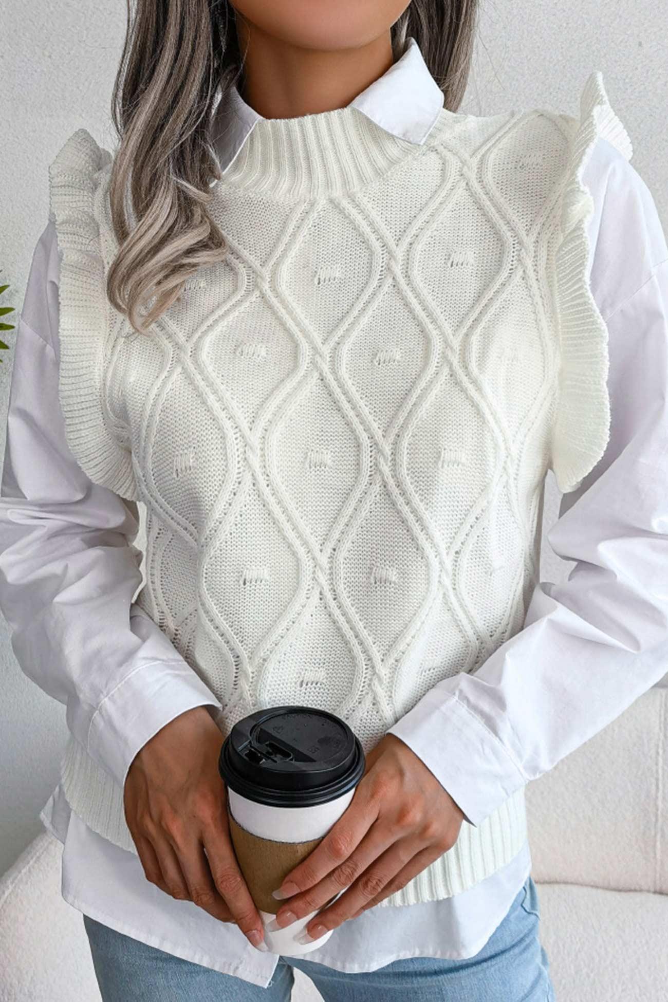 Woman holding a coffee cup while wearing a white sleeveless knit vest with a diamond pattern and ruffle trim over a white button-down shirt.