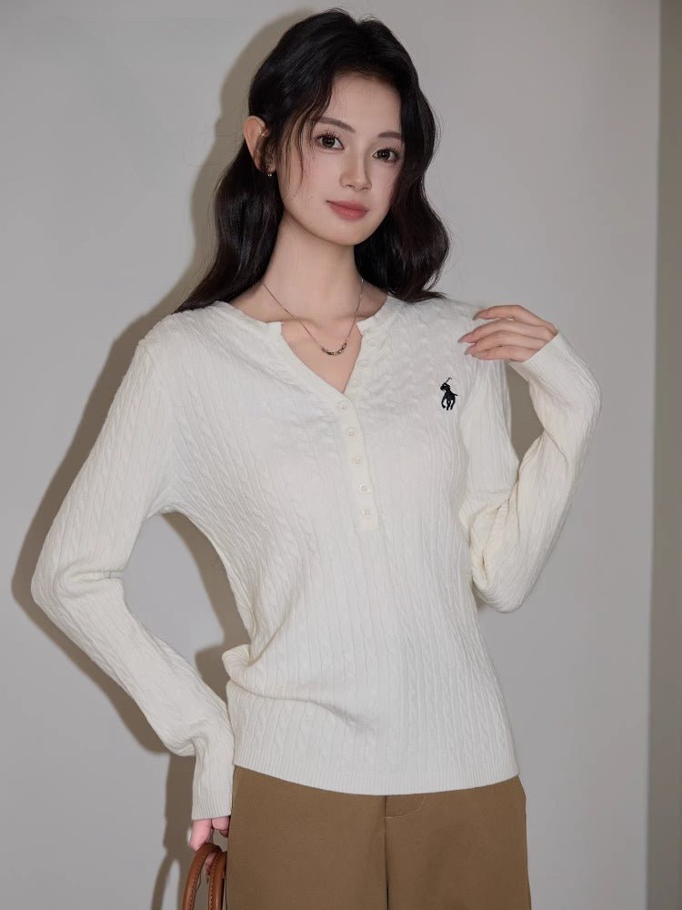 Woman wearing a cream cable knit long sleeve sweater with a V-neck and embroidered logo.