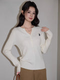 Woman wearing a cream cable knit long sleeve sweater with a V-neck and embroidered logo.