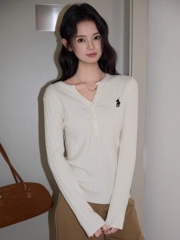 Woman wearing a cream cable knit long sleeve sweater with a V-neck and embroidered logo.