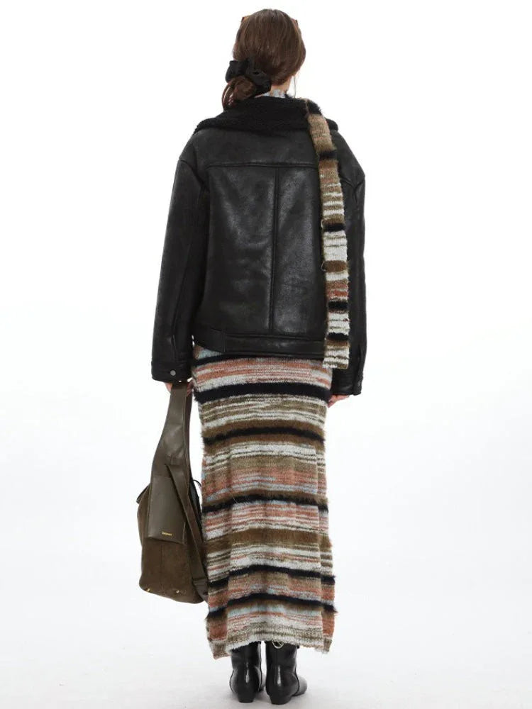 Back view of a woman in a black faux leather aviator jacket, striped maxi skirt, and olive green shoulder bag.