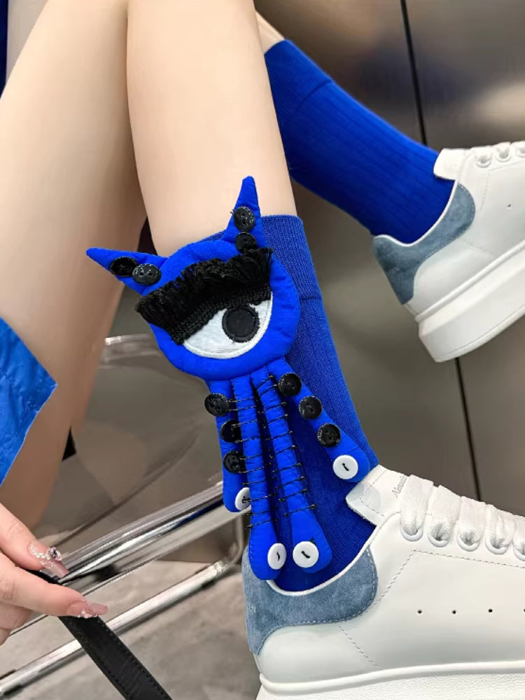 Close-up of blue novelty socks with an eye applique and button details on a person's legs, paired with white sneakers.