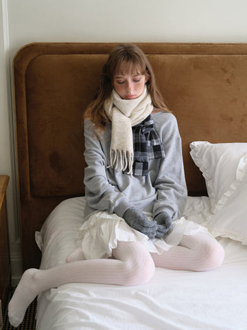 Woman wearing a grey sweatshirt, white ruffled skirt, light pink ribbed tights, and a cream scarf with fringe detail
