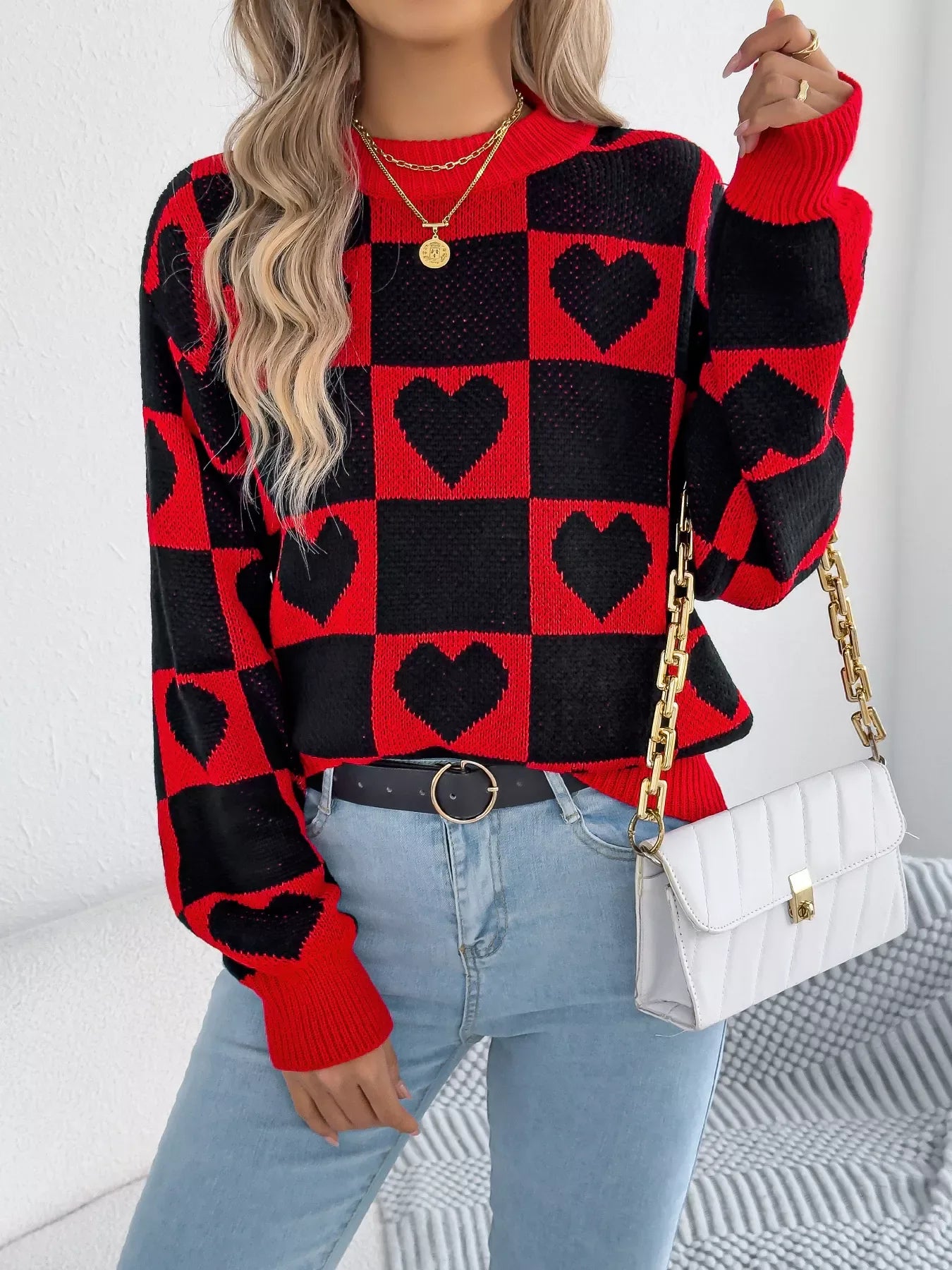 Woman wearing a red and black checkerboard knit sweater with black heart patterns, light wash denim jeans, and a white quilted handbag with a gold chain strap.