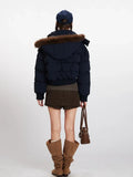 Back view of a woman in a navy blue cropped puffer jacket with a fur-trimmed hood, dark brown skirt, and brown boots.