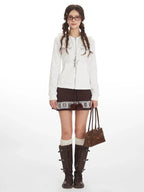 Woman wearing a white hooded zip-up jacket with a bow and heart graphic print, paired with a brown patterned skirt, white knee-high socks, and brown boots.