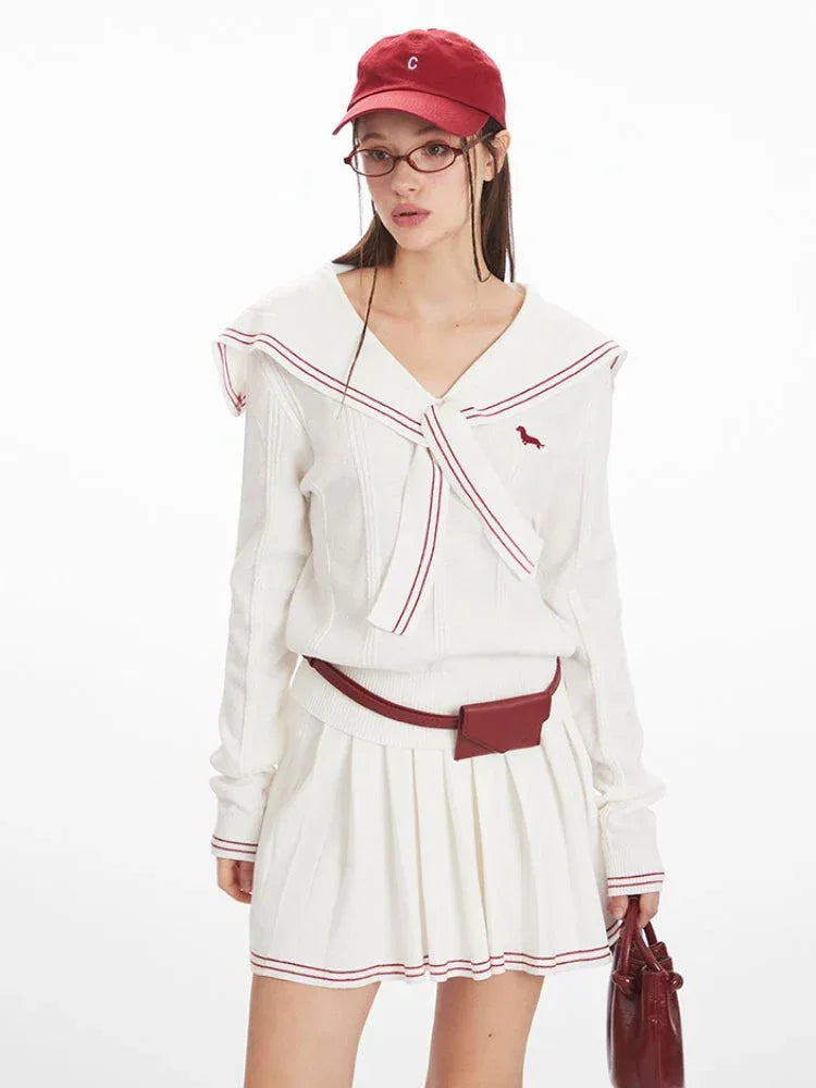 Front view of a woman wearing a white knit sweater with a navy sailor collar and red trim, a red belt bag, and a white pleated skirt.