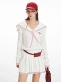 Front view of a woman wearing a white knit sweater with a navy sailor collar and red trim, a red belt bag, and a white pleated skirt.