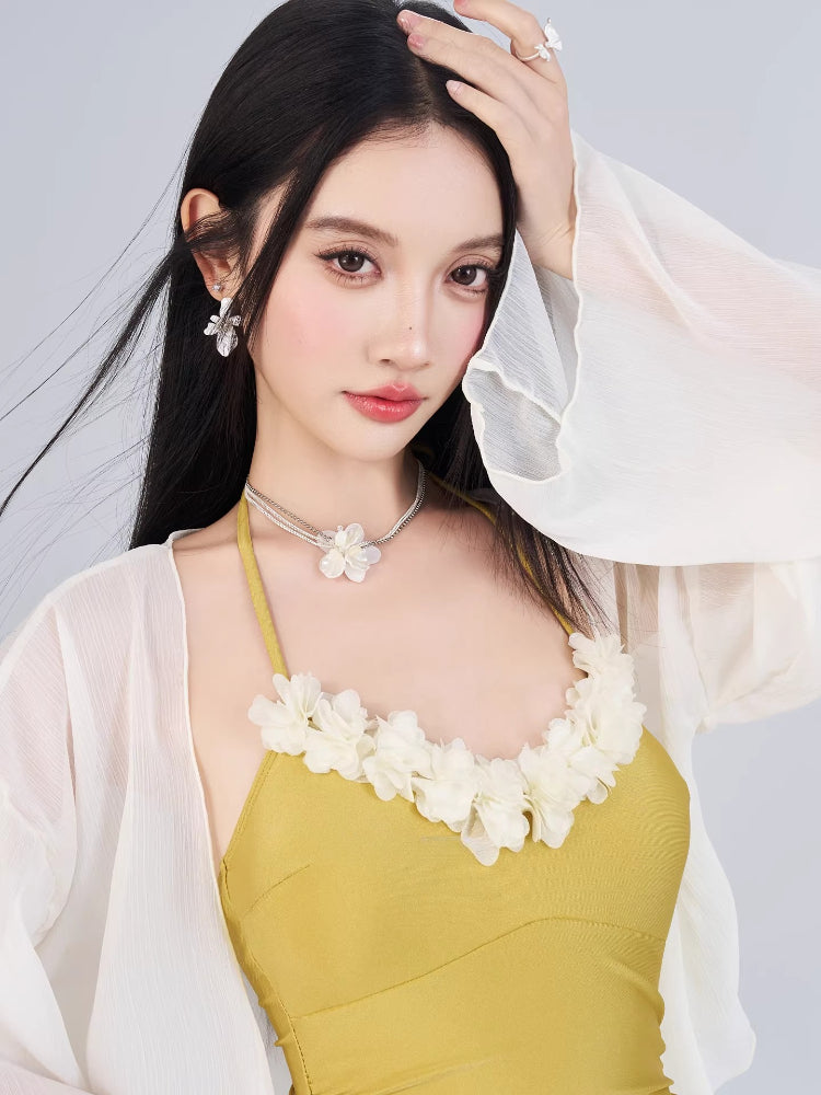 Woman wearing a mustard yellow one-piece swimsuit with floral trim and ruffled shorts, paired with a sheer white cover-up.