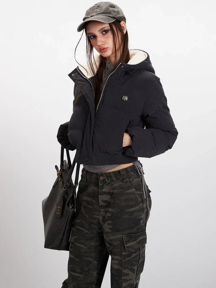 Close-up of a woman wearing a black cropped puffer jacket with a hood, grey t-shirt, and camouflage cargo pants, holding a black tote bag.