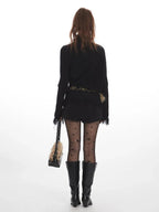 Back view of a woman wearing a black turtleneck sweater with fringed cuffs and hem, black shorts, black patterned tights, and black boots