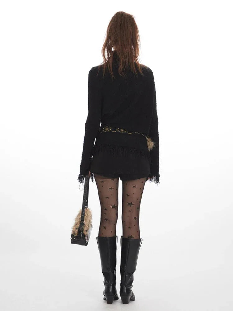 Back view of a woman wearing a black turtleneck sweater with fringed cuffs and hem, black shorts, black patterned tights, and black boots