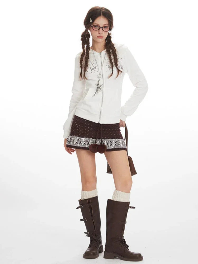 Full-body shot of a woman in a white hooded zip-up jacket, brown patterned skirt, white knee-high socks, and brown boots, holding a brown handbag.