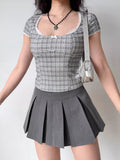 Woman wearing a grey plaid fitted top and a grey pleated skirt, accessorized with a silver handbag and black beaded necklace.