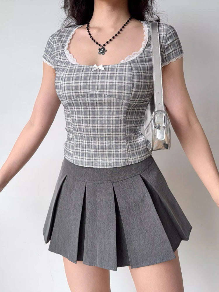 Woman wearing a grey plaid fitted top and a grey pleated skirt, accessorized with a silver handbag and black beaded necklace.