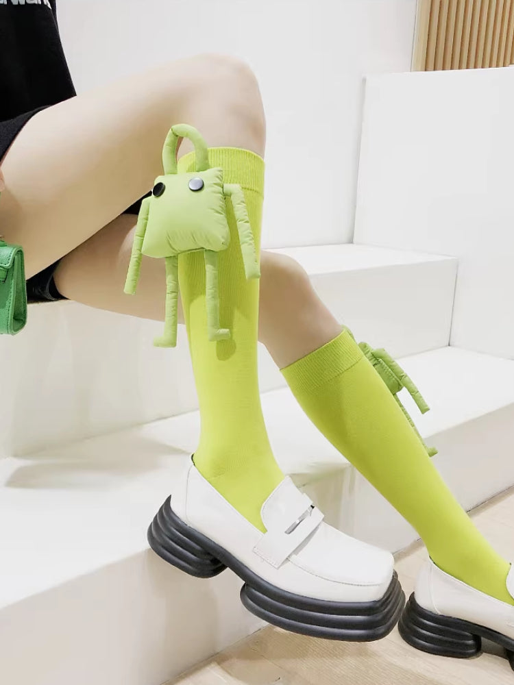 Lime green knee-high socks with a detachable plush frog toy on the right leg, paired with white loafers.