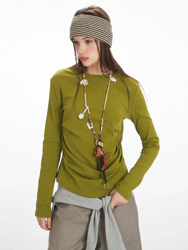 Close-up of a model wearing a long-sleeve olive green top with ruched side detail, grey layered vest, taupe wide-leg pants, yellow handbag, and striped headband.