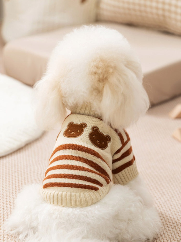 The back of a small white dog wearing a cream and brown striped knit sweater with two embroidered bear faces on the upper back.