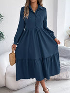 Full-length view of a woman in a navy blue midi dress with a collared neckline and tiered skirt, holding a straw bag.