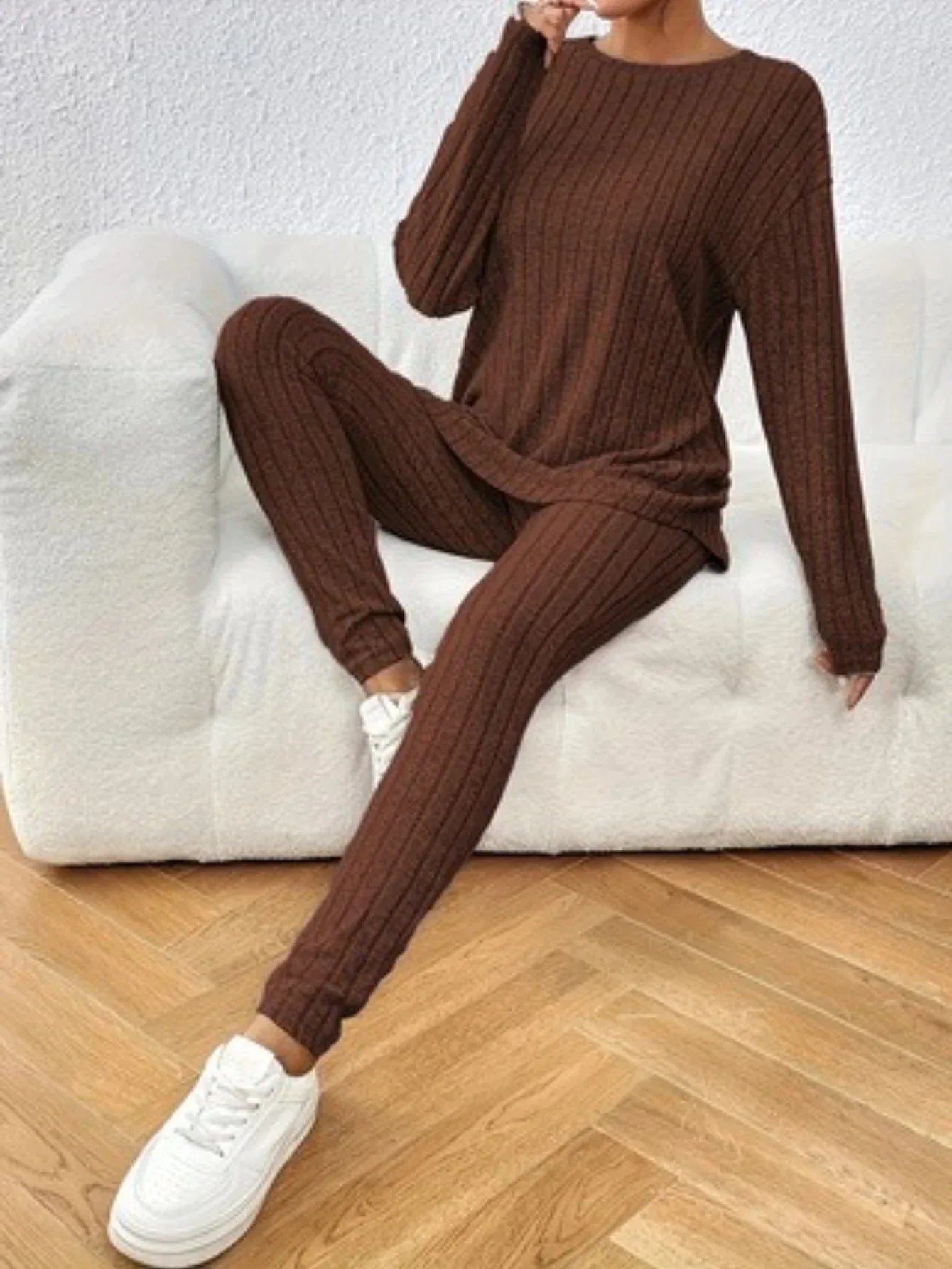 Woman wearing a brown ribbed knit loungewear set with a crew neck top and slim-fit pants, sitting on a white couch.