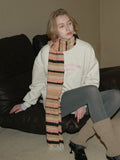 Woman wearing a cream-colored sweatshirt, grey leggings, leg warmers, a multi-colored striped scarf, and black loafers sitting on a black couch, looking forward.