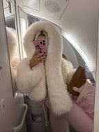 Woman taking a mirror selfie on an airplane wearing a cream-colored faux fur cropped jacket with an oversized hood and pink sweatpants