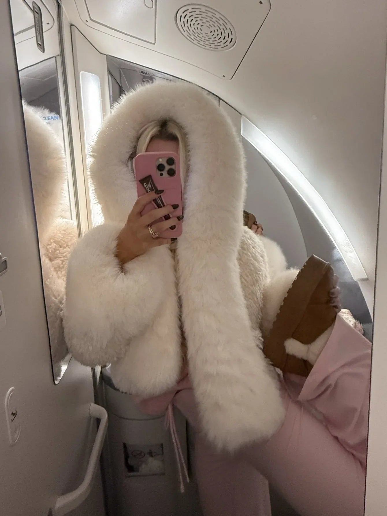 Woman taking a mirror selfie on an airplane wearing a cream-colored faux fur cropped jacket with an oversized hood and pink sweatpants