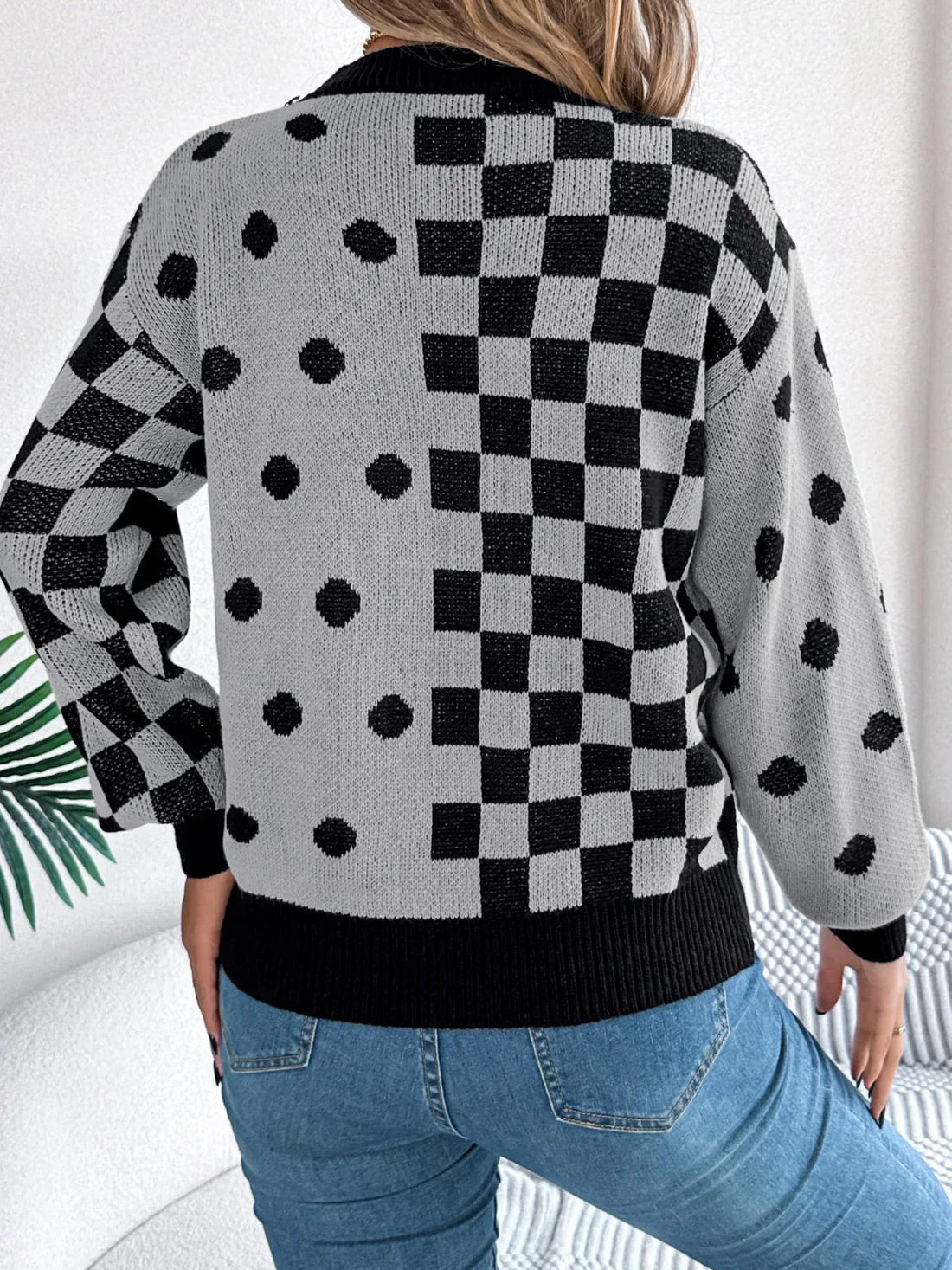 Woman wearing a grey knit sweater with a black checkerboard and polka dot pattern, paired with blue jeans and a white handbag.