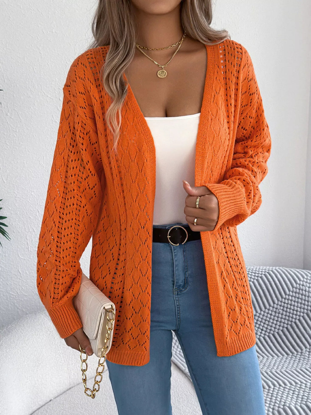 Woman holding a white clutch bag while wearing an orange open-knit cardigan.
