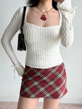 Woman wearing a white textured long sleeve top and a red plaid skirt, holding a black handbag.