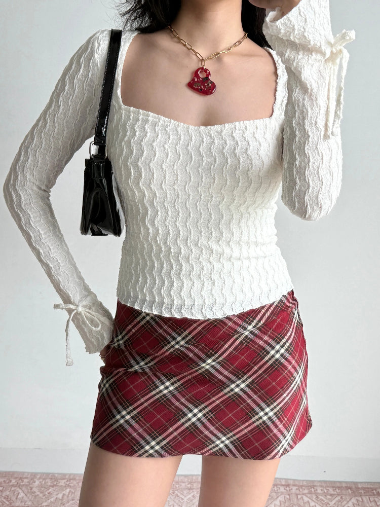 Woman wearing a white textured long sleeve top and a red plaid skirt, holding a black handbag.