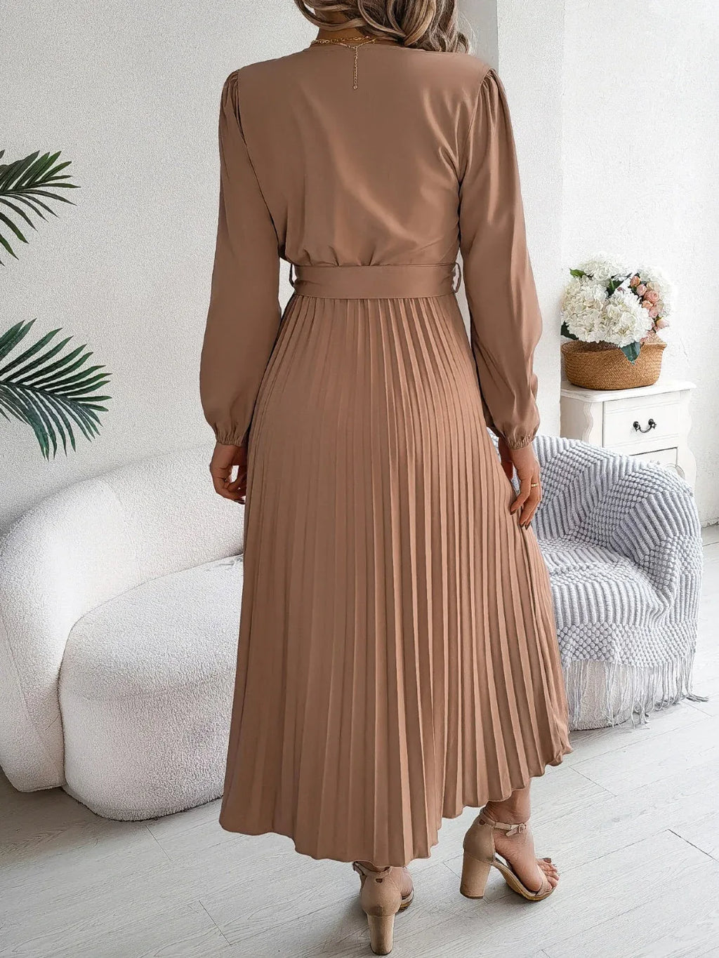Back view of a woman in a brown long-sleeve V-neck midi dress with a pleated skirt and a self-tie belt