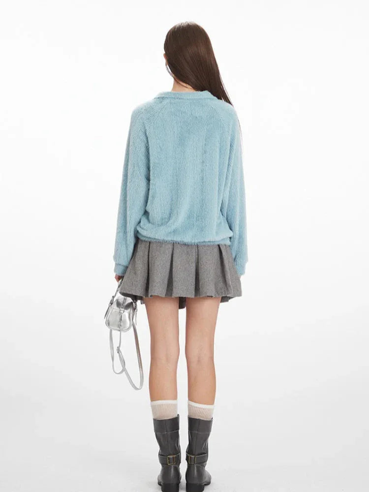 Back view of a model wearing a light blue fuzzy knit long-sleeve collared v-neck sweater with a ribbed hem, paired with a grey pleated mini skirt, grey boots, and a silver handbag.