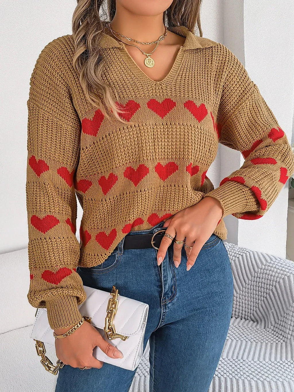 Woman in a tan knit sweater with a red heart pattern, blue jeans, and a white handbag.