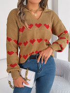 Woman in a tan knit sweater with a red heart pattern, blue jeans, and a white handbag.