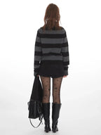 Rear view of a woman wearing a grey and black striped knit sweater with a cowl neck, black shorts, black patterned tights, and black boots, holding a black handbag.