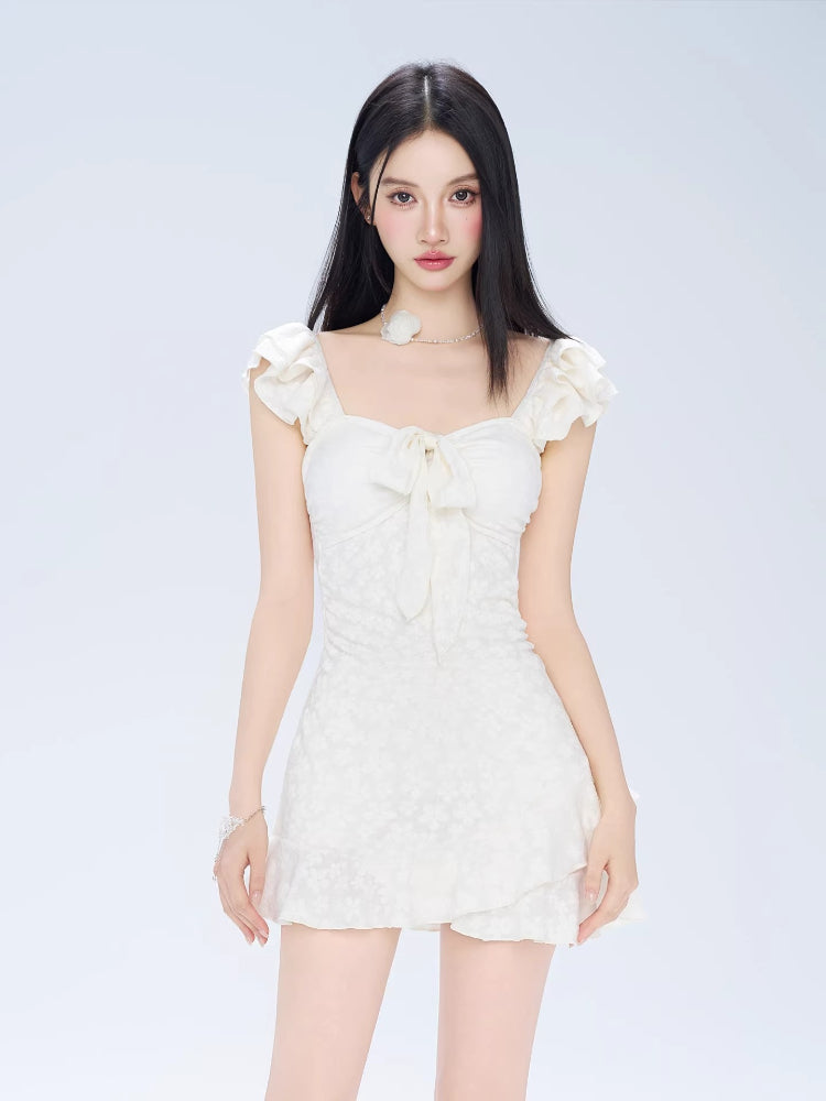 Woman wearing a white jacquard fit and flare mini dress with ruffled cap sleeves and a sweetheart neckline with a bow detail.
