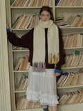 Woman leaning against a bookshelf while wearing a cream scarf with ombre fringe.