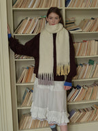 Woman leaning against a bookshelf while wearing a cream scarf with ombre fringe.