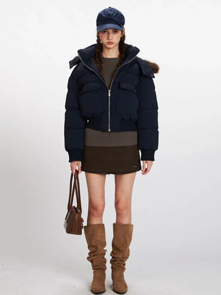 Woman wearing a navy blue cropped puffer jacket with a fur-trimmed hood, dark brown skirt, and brown boots, holding a brown handbag.