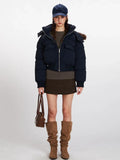 Woman wearing a navy blue cropped puffer jacket with a fur-trimmed hood, dark brown skirt, and brown boots, holding a brown handbag.