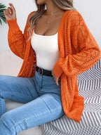 Woman wearing an orange open-knit cardigan with long sleeves over a white tank top and blue jeans.