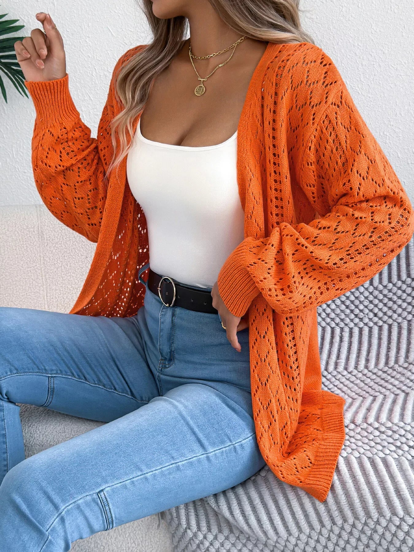 Woman wearing an orange open-knit cardigan with long sleeves over a white tank top and blue jeans.